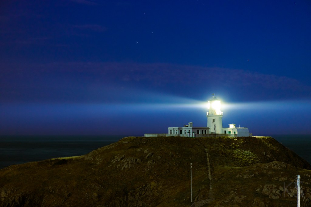 Strumble Head lighthouse casting an intense beam of light against a deep blue sky.